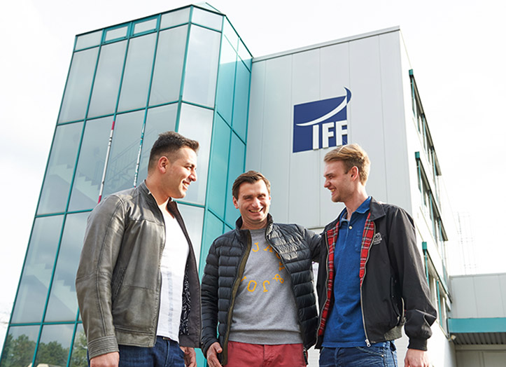 Three men stand smiling and talking outside a modern glass building with the IFF logo on the wall. The sky is bright and the mood is friendly and casual.