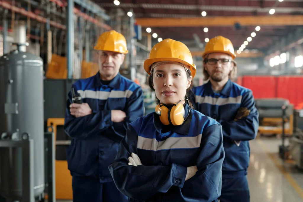 Three factory workers in blue uniforms and yellow hard hats stand with arms crossed in an industrial setting, with one person in the foreground wearing ear protection around their neck.